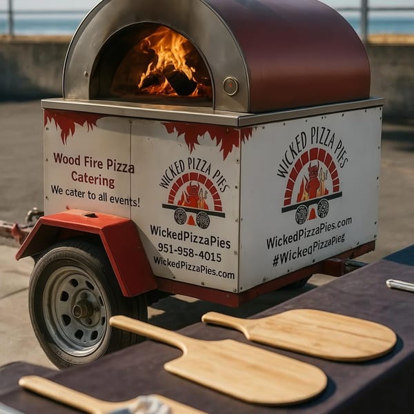 Wicked Pizza Pies mobile wood-fired oven with fire burning, and pizza peels on a table outdoors.