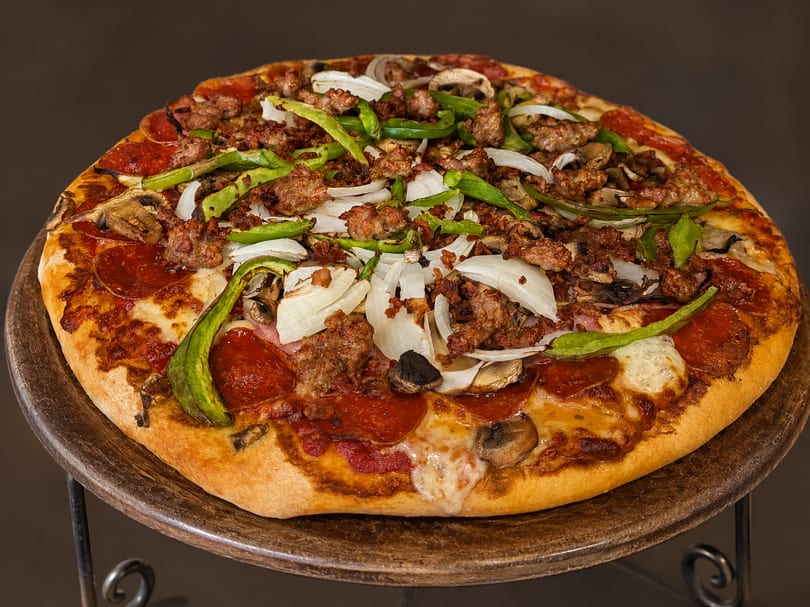 Loaded pizza with pepperoni, sausage, green peppers, onions, and mushrooms on a metal stand.