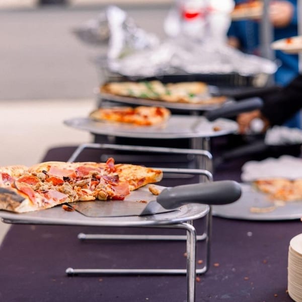 Guests serving themselves from a buffet line of various pizzas on metal stands.