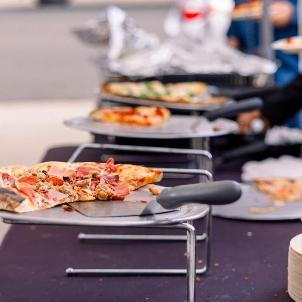 Guests serving themselves from a buffet line of various pizzas on metal stands.