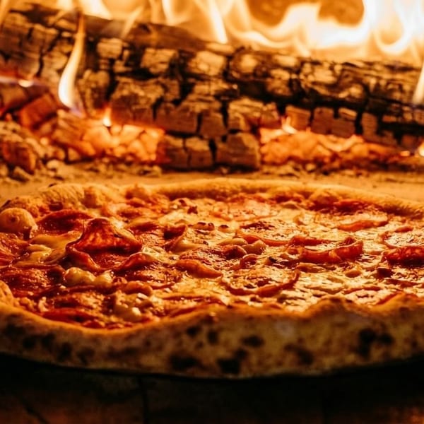 Pepperoni pizza baking in a glowing wood-fired oven with bright flames.