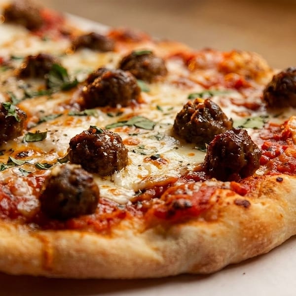 Close-up of a meatball pizza with melted mozzarella, red sauce, and fresh basil on a white surface.