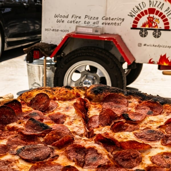 A freshly baked pepperoni pizza on a wooden peel in front of a "Wicked Pizza Pies" catering trailer.