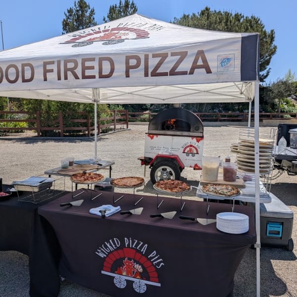 Wicked Pizza Pies mobile wood-fired pizza setup with multiple pizzas displayed under a tent outdoors.